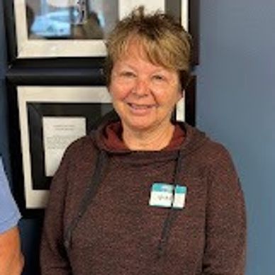 Smiling woman in a brown hoodie with a name tag stands indoors against a blue and white framed wall.