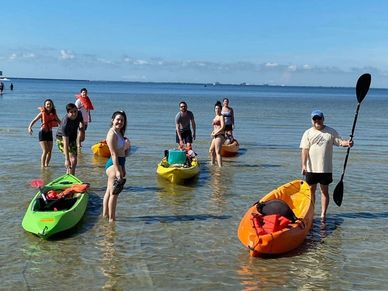 8 kayakers standing in shallow water with kayaks. blue sky in background www.whatsuppaddlesports.com