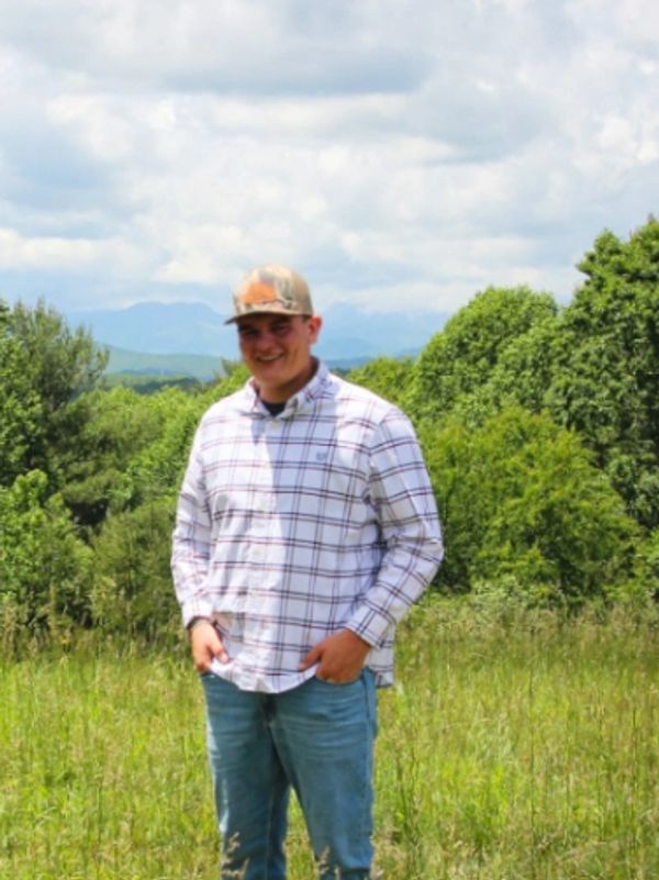 A young man in a plaid shirt and cap stands in a grassy field with trees and mountains in the background.