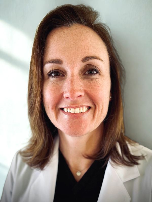 Smiling woman in a white coat, likely a healthcare professional, with light makeup and a friendly expression.