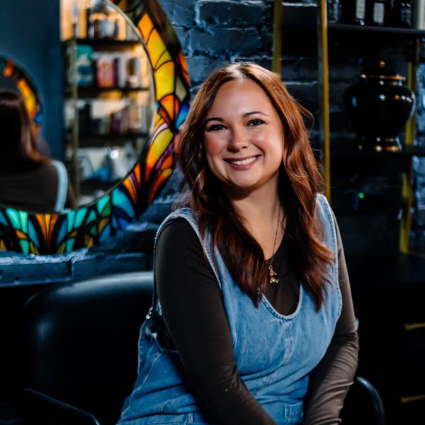 Smiling woman with auburn hair sitting indoors by a colorful mirror.