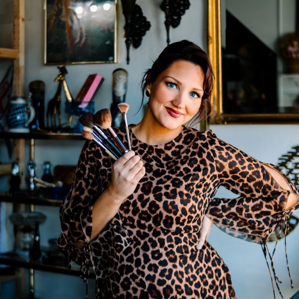 Woman in leopard print dress holding makeup brushes smiling.