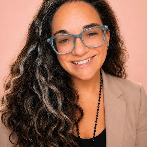Smiling woman with long curly hair and large glasses against a peach background.