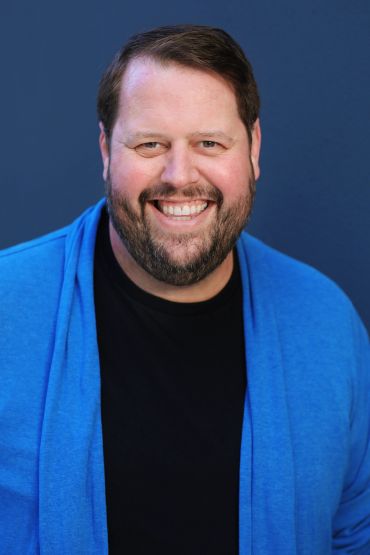 Smiling man in black shirt and blue cardigan.