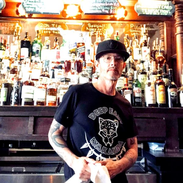 Bartender in a black cap and Wakanda shirt stands in front of a well-stocked bar.