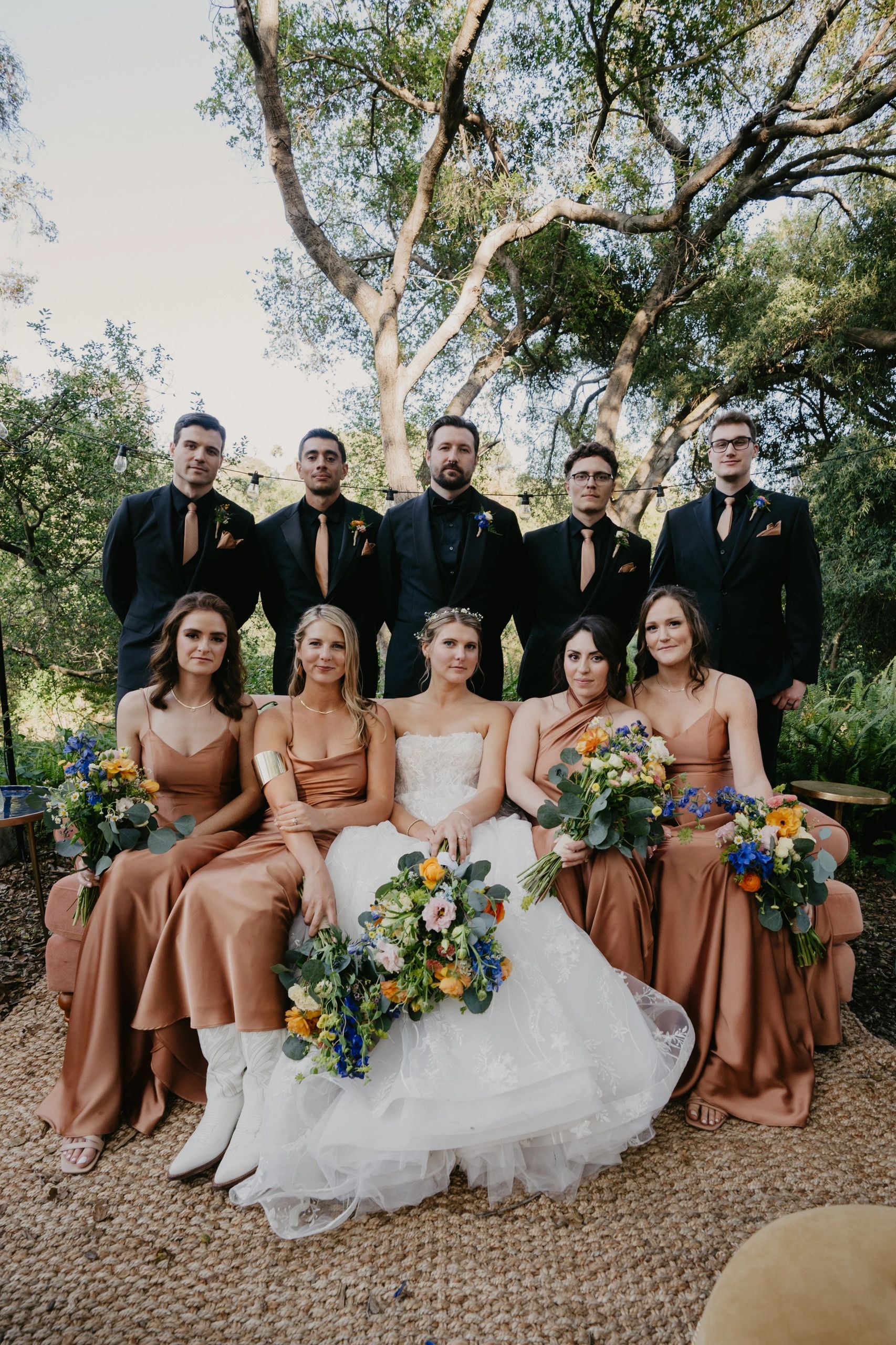Wedding party with bride, bridesmaids in copper dresses, and groomsmen in black suits outdoors.