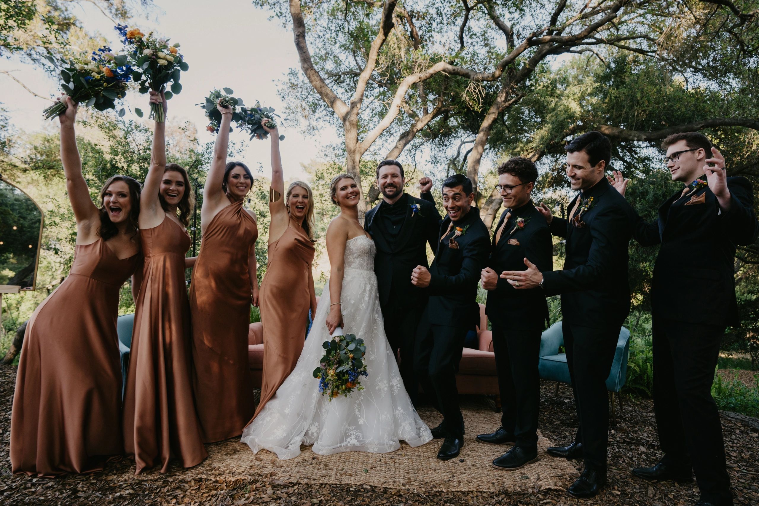 Joyful wedding party celebrates outdoors with bridesmaids in bronze dresses and groomsmen in black suits.