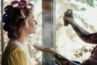 A woman with hair rollers gets hairspray applied by a tattooed stylist.
