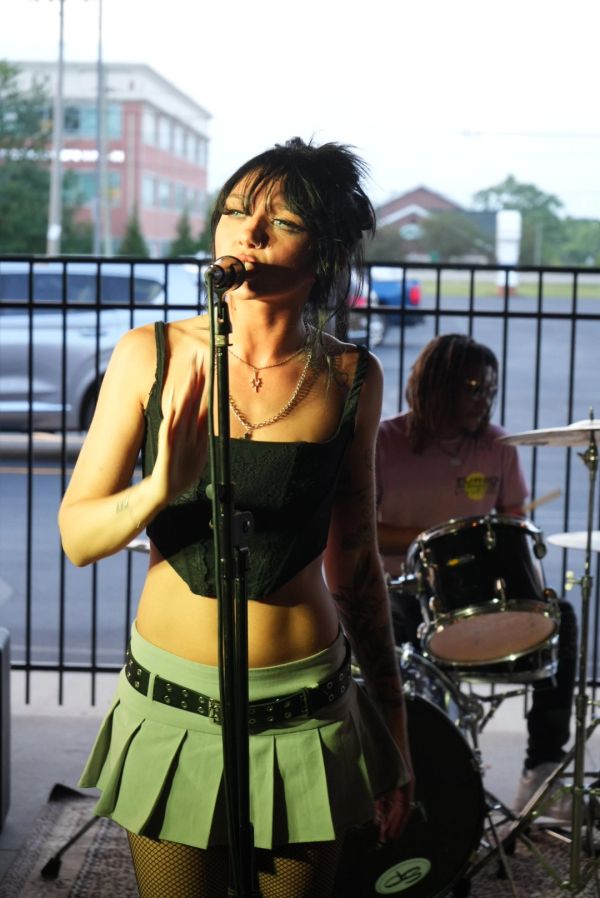 Woman singing into microphone with drummer in background.