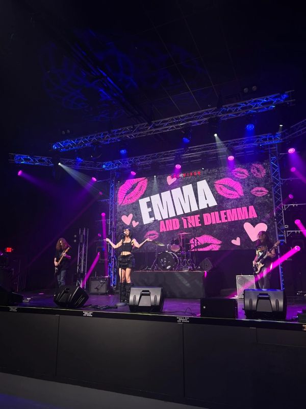 Band performing under bright lights on stage with 'EMMA AND THE DILEMMA' on screen.