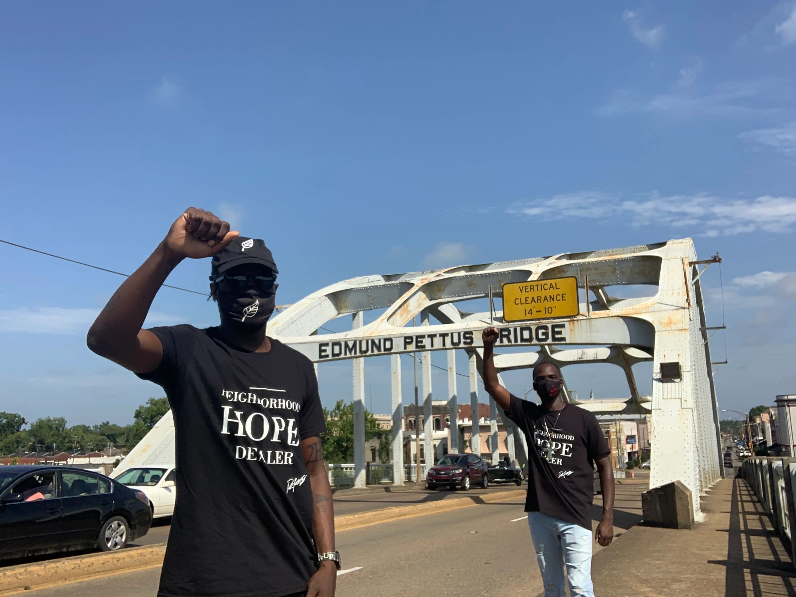 Two men raising fists on Edmund Pettus Bridge, wearing matching shirts and masks.