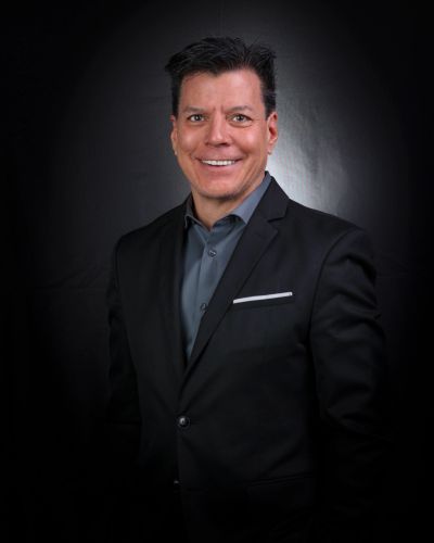 Man in a black suit smiling against a dark background.