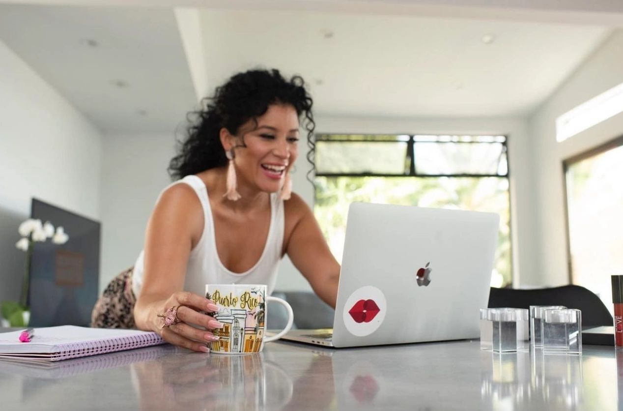 A cheerful woman with curly hair working on a laptop while holding a Puerto Rico mug.