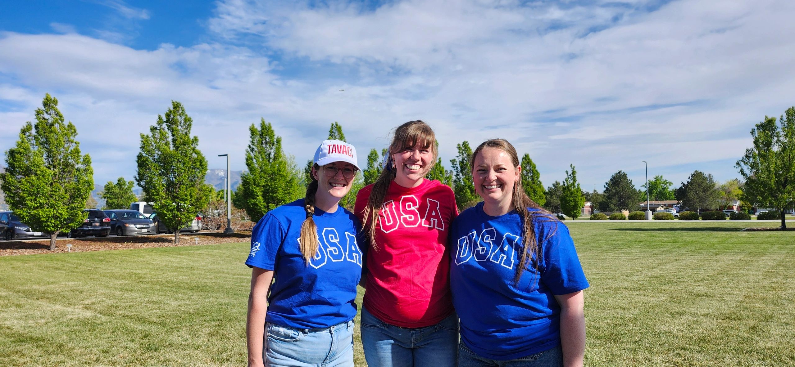 Three women smiling outdoors wearing USA shirts in red and blue.