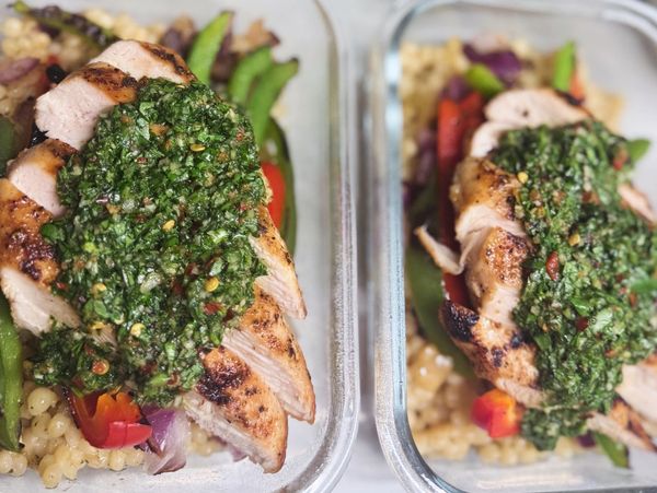Grilled chicken with chimichurri sauce over couscous and vegetables in meal prep containers.
