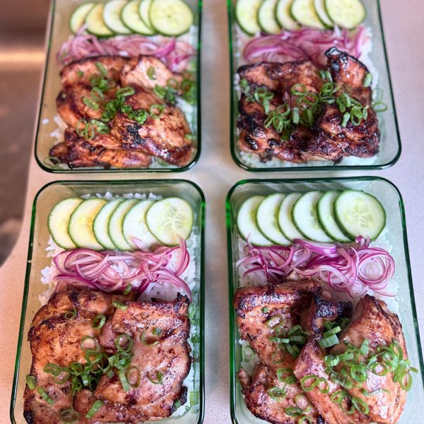 Four meal prep containers with grilled chicken, rice, cucumber, and pickled onions.