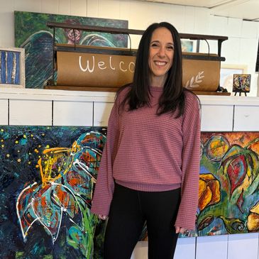 A smiling woman stands in front of colorful paintings and a welcome sign.