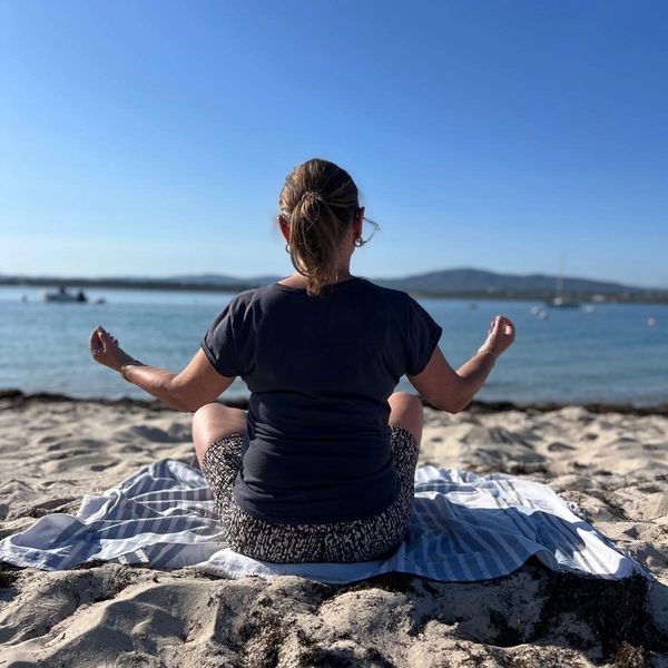 A woman meditates on a striped towel by the beach with a calm sea and clear sky.