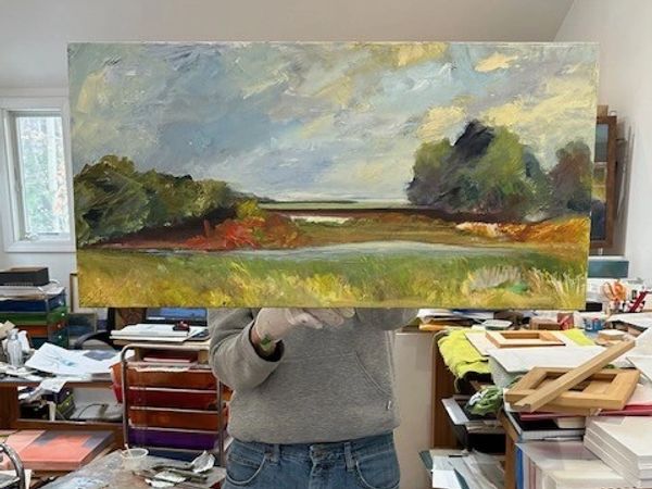 Person holding a landscape painting in an art studio.