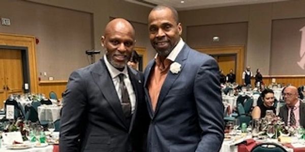 Two men in suits posing at a formal event with decorated tables in the background.