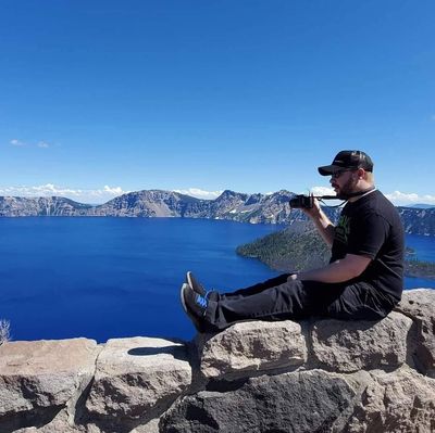 Person taking photo by blue lake from rocky ledge.