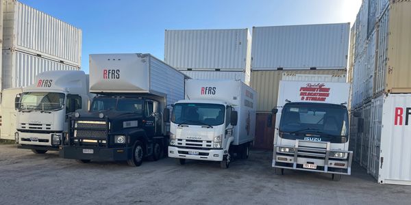 Four trucks parked in front of stacked shipping containers under clear sky.