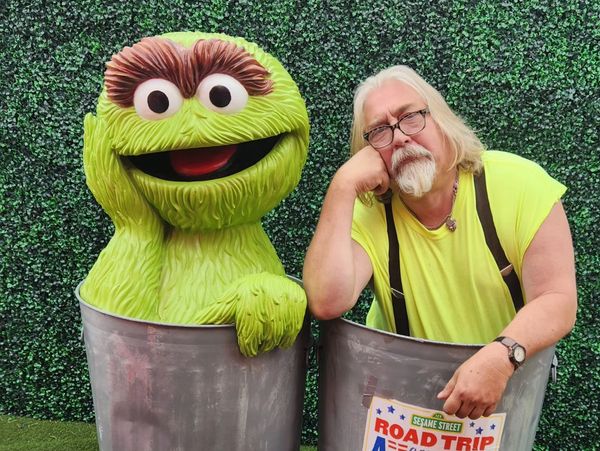 Man and Oscar the Grouch in trash cans with green hedge background.