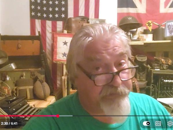 Older man with glasses in a room filled with vintage military items and flags.