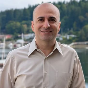 Smiling man standing outdoors with greenery and water in the background.