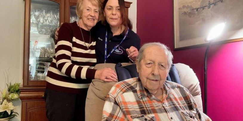 Three people posing indoors, an elderly man seated and two women standing behind him.