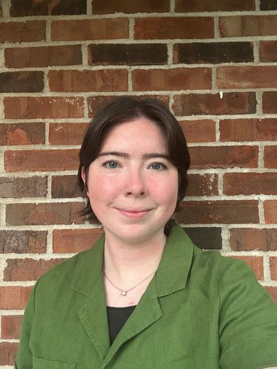 Smiling person in a green jacket against a brick wall.