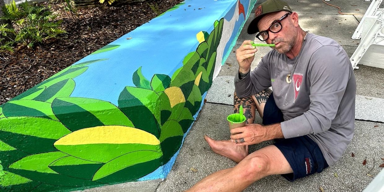 David Wirth seated beside a colorful tropical mural while painting bright green foliage.