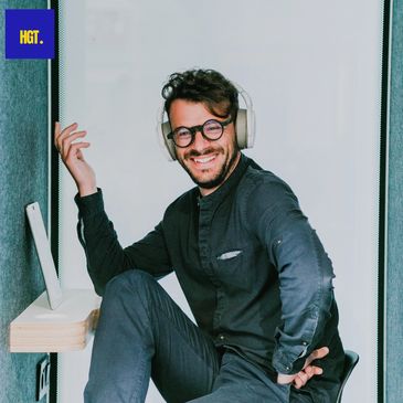 A bearded man with glasses and headphones, smiles while sitting in an office with a tablet.