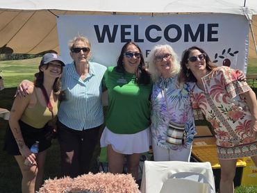 Five women smiling under a welcome banner at an outdoor event.