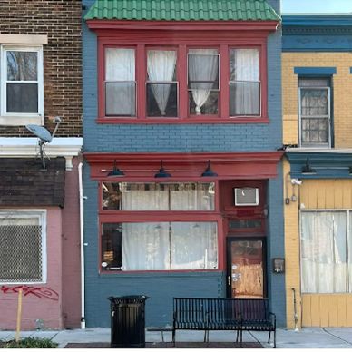 Small blue storefront with red trim and green roof, flanked by colorful buildings.
