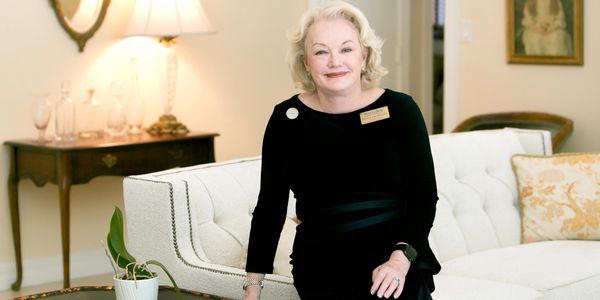 Elderly woman in black dress smiling while sitting on a white sofa in a well-lit room.