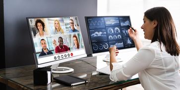 A woman in front of two computer screens speaking on a video call with her six colleagues 