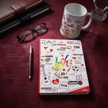 Desk setup with a colorful notebook, glasses, pen, and a mug on a maroon surface.