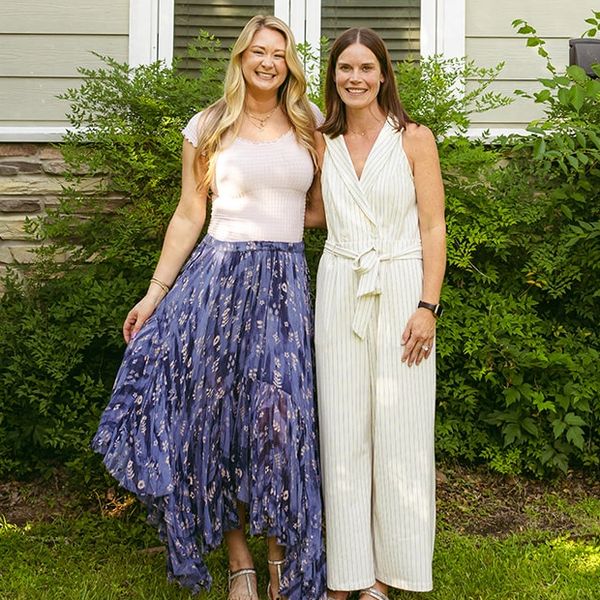 Two women smiling outdoors in front of greenery.