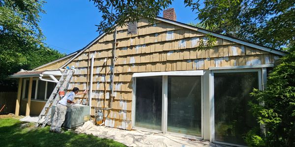 A person patching and painting the exterior siding of a house on a sunny day.