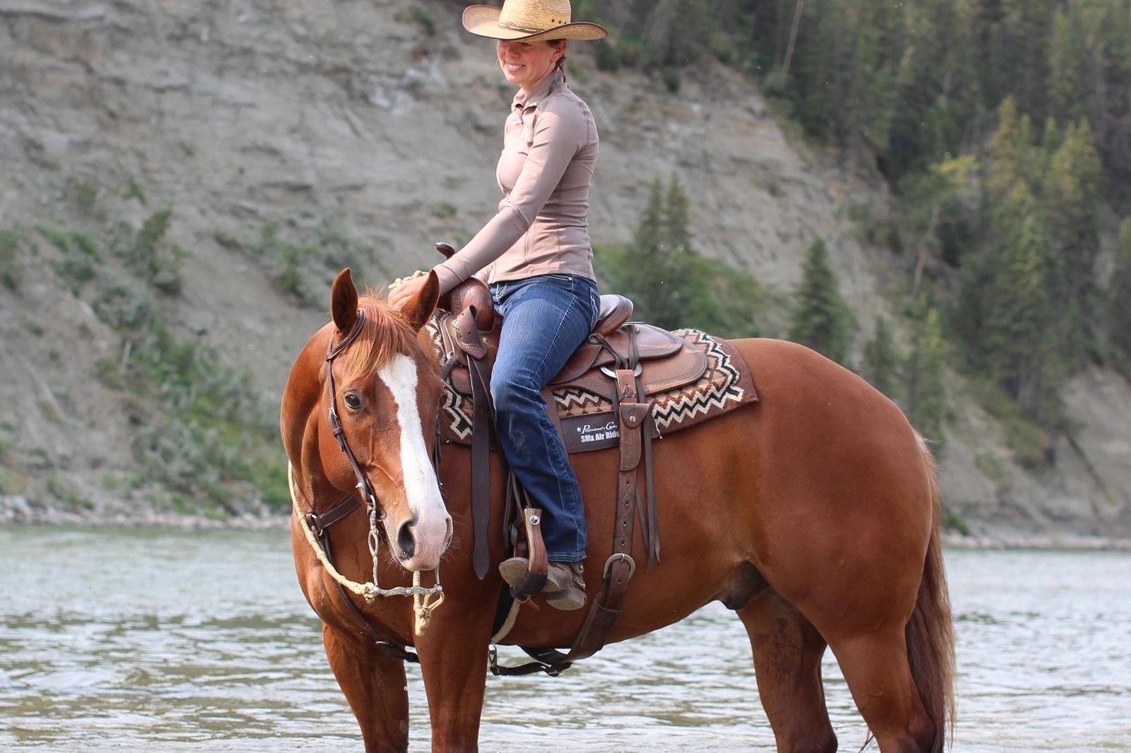 Woman wearing a cowboy hat riding a horse by the river.