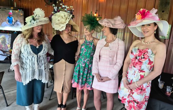 Five women dressed in elegant attire with large floral hats at a social event.