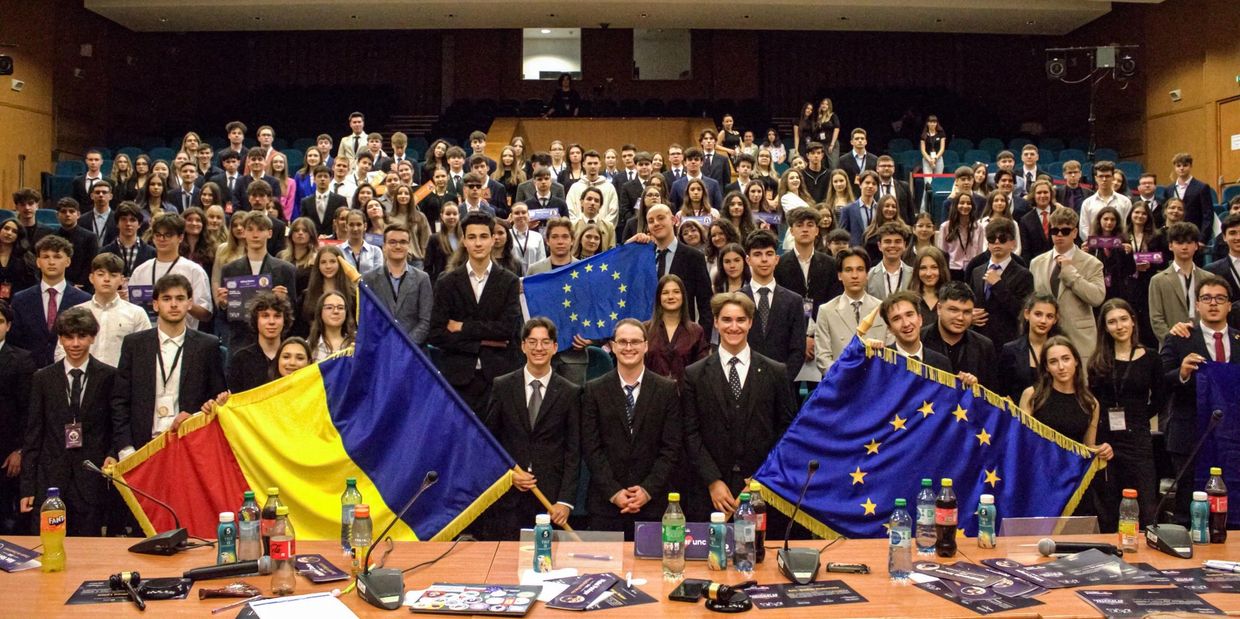 A large group of young people posing with European Union and Romanian flags in a conference room.