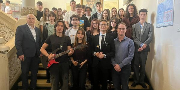 A diverse group of young people and adults posing on stairs, some holding a guitar and a gavel.