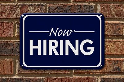 A 'Now Hiring' sign on a brick wall.