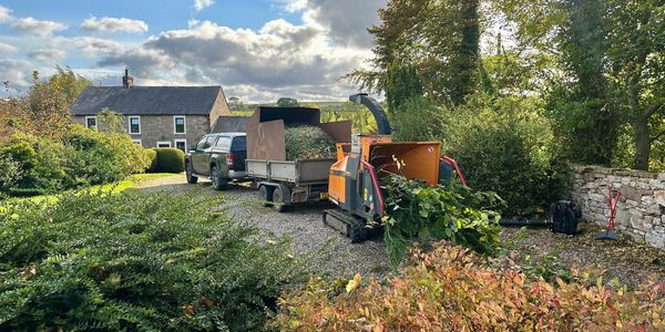 Wood chipper beside a trailer loaded with garden waste near a rural house.