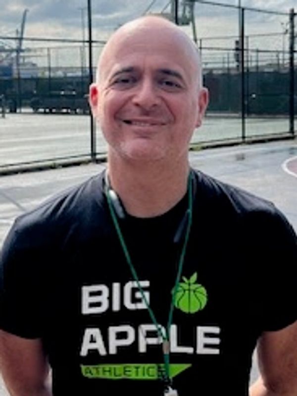 Man smiling in a basketball court with a "Big Apple Athletic" shirt.