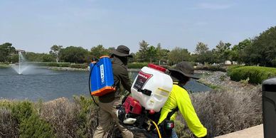 Dos personas fumigando cerca de un lago en un día soleado.
