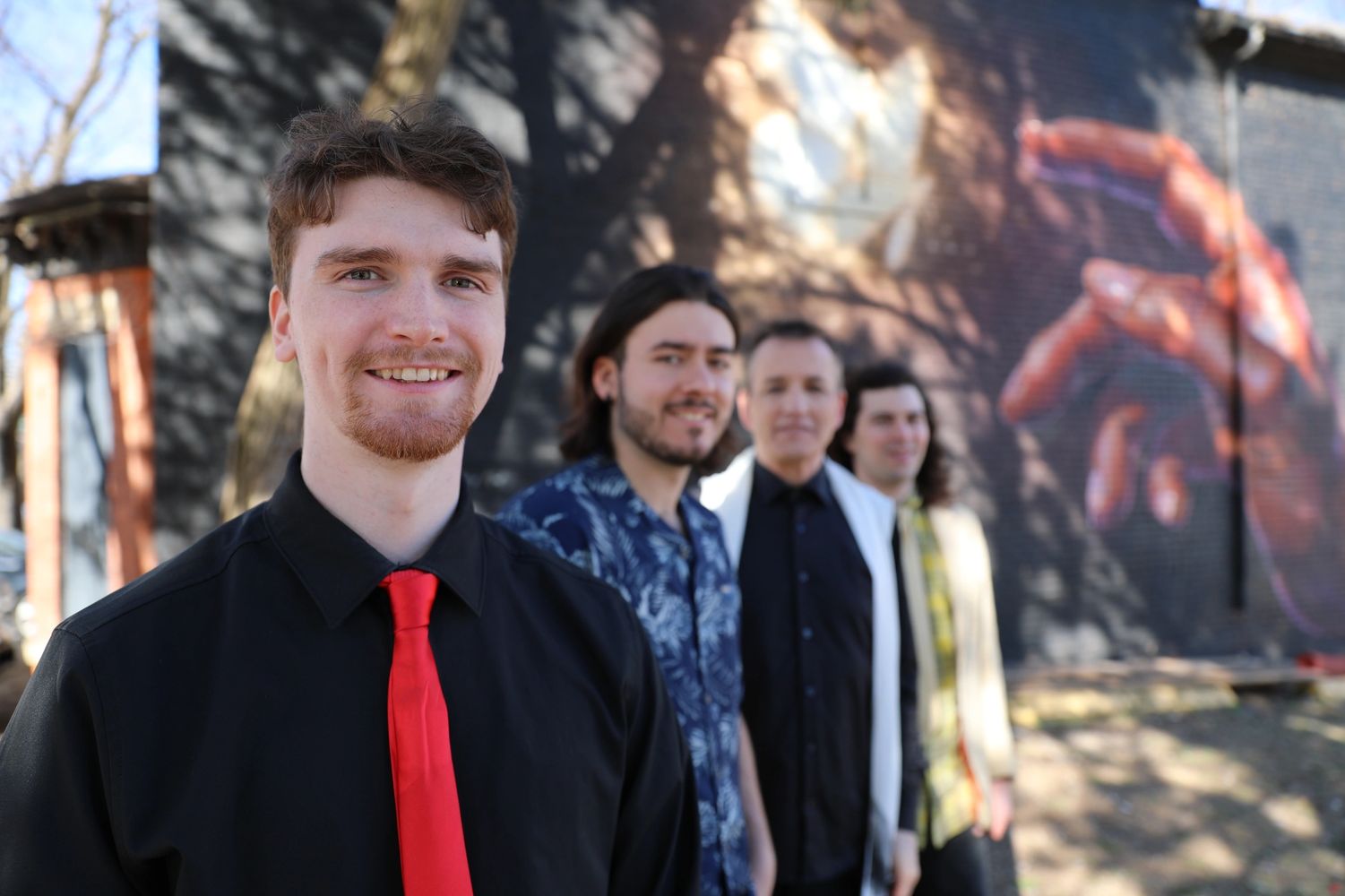 Four men standing in a line outdoors with a mural of a hand on the wall behind them.
