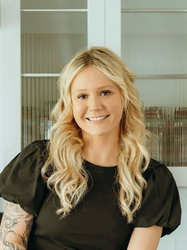 Smiling woman with blonde curly hair and tattooed arm in black top.